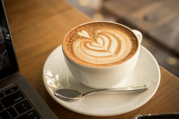 カップマグカップやノートパソコンのラテアートミルク泡とホットコーヒーラテのクローズアップや カフェのコーヒーショップの木の机の上のスマートフォン ビジネスコンセプト中 — ストック写真
