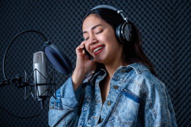 Profesyonel bir stüdyoda mikrofon önünde şarkı kaydeden, kulaklık takan genç Asyalı kadın vokalistin mutlu bir gülümsemesi.