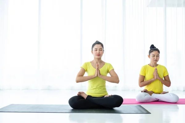 Two young Asian women workout practicing yoga in yellow dress or pose with a trainer and practice meditation wellness lifestyle and health fitness concept in a gym.