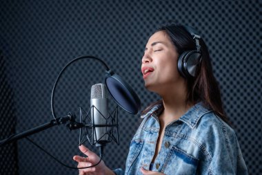 Profesyonel bir stüdyoda mikrofon önünde şarkı kaydeden, kulaklık takan genç Asyalı kadın vokalistin mutlu bir gülümsemesi.