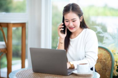 Asyalı serbest çalışanlardan memnun. İş kadını, kafeteryada bilgisayarla ve kahve bardağıyla iş hayatı tarzı iletişim konseptiyle günlük akıllı telefon konuşmaları yapıyor.