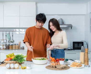 Sevgi dolu genç bir çiftin mutlu portresi. Asyalı neşeli bir şekilde yemek pişirmeyi seviyor. Sebze, et, ekmek pişiriyor.