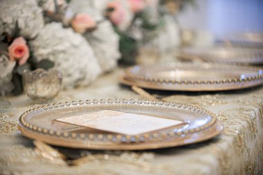 Muhteşem masa manzarası altın şarj tabakları, davetiye ve çiçekler düğün resepsiyonunda altın desenli masa örtüsü üzerine dizilmişti.