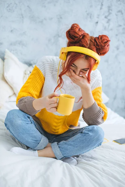 Bir kız yatağa oturur, kahve içer, güler ve sarı kulaklıkla müzik dinler..