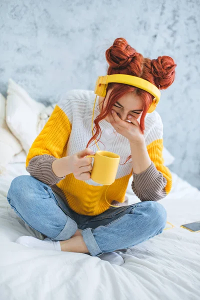 Дівчина сидить на ліжку, п'є каву, сміється і слухає музику в жовтих навушниках . — стокове фото