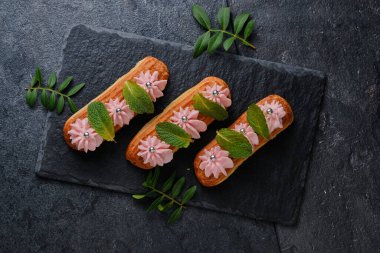 Nane yapraklarıyla süslenmiş pembe kremalı ekler. Siyah bir tabakta tatlı. Koyu arkaplanda kekler.