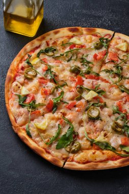 Pizza with tomatoes, shrimps, arugula, olives, lemon, mozzarella cheese, spices and tomato sauce. On a dark background.