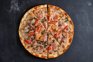 Pizza with tomatoes, ham, capers, mushrooms, mozzarella cheese, spices, sesame seeds and tomato sauce. Flat lay. On a dark background.