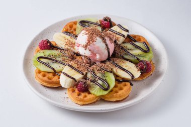Sweet Belgian waffles for breakfast on a light plate. Waffles with ice cream, chocolate, kiwi, banana and raspberries. Dish on a white plate.