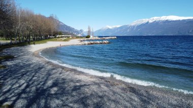Toscolano Maderno 'daki çakıl taşı plajı. Karlı dağları ve Garda Gölü, İtalya.