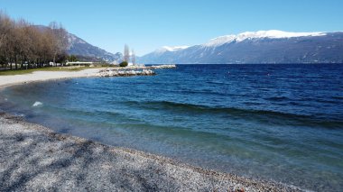 Toscolano Maderno 'daki çakıl taşı plajı. Karlı dağları ve Garda Gölü, İtalya.