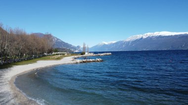 Toscolano Maderno 'daki çakıl taşı plajı. Karlı dağları ve Garda Gölü, İtalya.