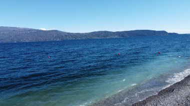 Toscolano Maderno 'daki çakıl taşı plajı. Karlı dağları ve Garda Gölü, İtalya.
