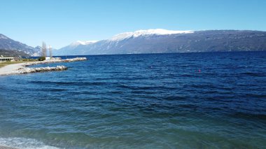 Toscolano Maderno 'daki çakıl taşı plajı. Karlı dağları ve Garda Gölü, İtalya.