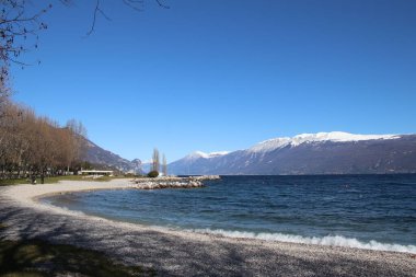 Toscolano Maderno 'daki çakıl taşı plajı. Karlı dağları ve Garda Gölü, İtalya.