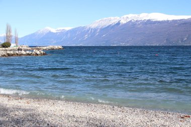 Toscolano Maderno 'daki çakıl taşı plajı. Karlı dağları ve Garda Gölü, İtalya.