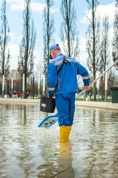 Un hombre en uniforme de trabajo azul limpia la superficie de la fuente en el parque con una red especial. Un trabajador del parque en uniforme lava el agua en la piscina. El servicio de parque elimina las hojas del embalse. — Foto de Stock