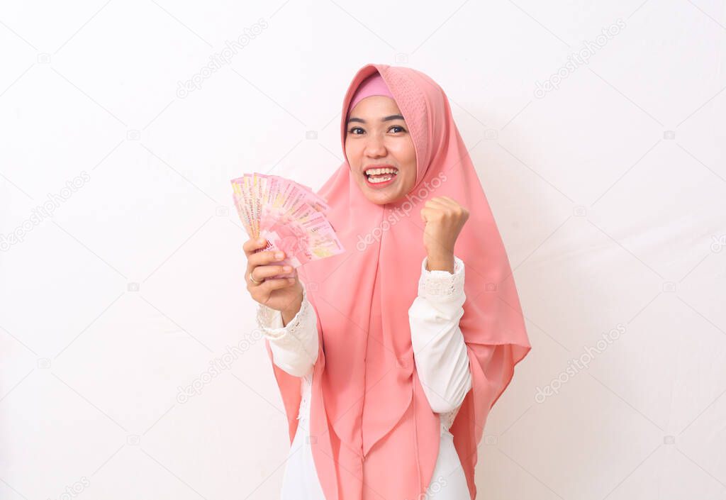 Happy asian woman in Muslim dress stands up, holding Indonesian money. Isolated on white background