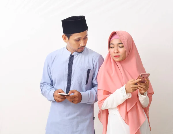 亚洲穆斯林夫妇站在孤立的背景下 用智能手机相互偷看对方的肖像 — 图库照片