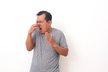 Asyalı bir adam beyaz arka planda izole edilmiş pis ve iğrenç bir koku alıyor. Nefesini burnunda tutuyor. Kötü koku konsepti.