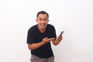 Siyah tişörtlü, cep telefonunu gösteren ve kameraya gülümseyen, cihaz veya mobil uygulama öneren tatmin olmuş Asyalı bir adamın portresi. Beyaz arka planda izole edilmiş kapalı stüdyo çekimi