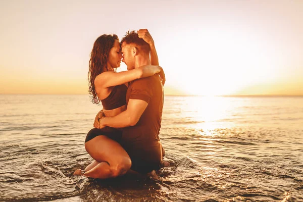 Happy young fit couple in sea or ocean hug each other with love at summer sunset. Romantic mood, tenderness, relationship, vacation concept.