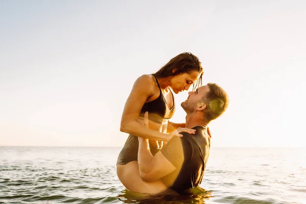 Happy young fit couple embrace each other with love in the sea or ocean at summer sunset. Romantic mood, tenderness, relationship, vacation concept.