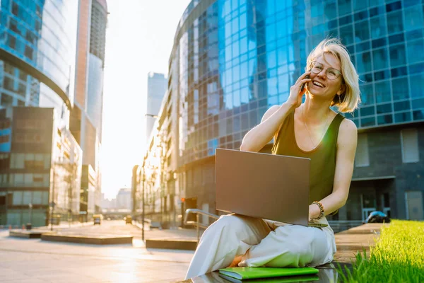 Blonde business woman sitting with laptop among the skyscrapers and speaks on the phone.