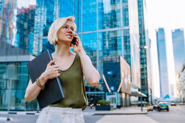 Young blonde business woman standing with laptop among the skyscrapers and speaks on the phone.