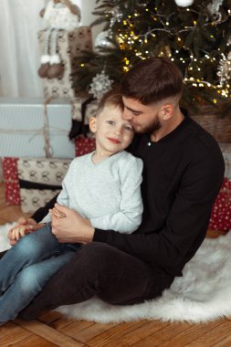 Kış tatili için süslü bir odada Noel ağacının yanında gülümseyen, sarılan ve oturan bir baba. Aile şenlik portreleri. Yeni yıl kutlamaları. Kış büyüsü zamanı, peri masalı heyecanı.
