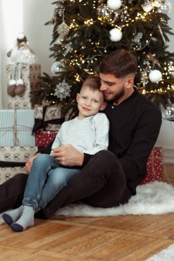 Kış tatili için süslü bir odada Noel ağacının yanında gülümseyen, sarılan ve oturan bir baba. Aile şenlik portreleri. Yeni yıl kutlamaları. Kış büyüsü zamanı, peri masalı heyecanı.