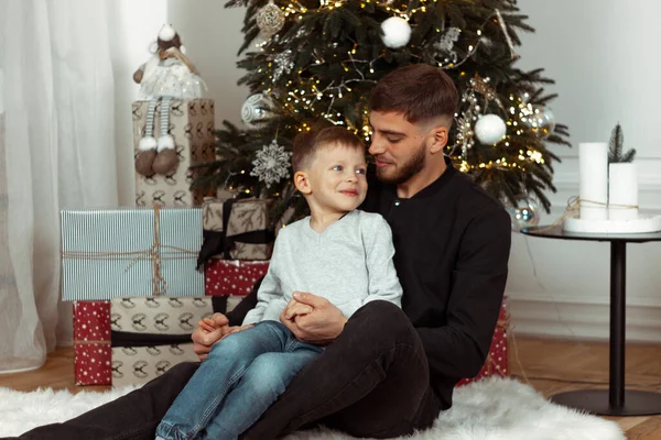 Kış tatili için süslü bir odada Noel ağacının yanında gülümseyen, sarılan ve oturan bir baba. Aile şenlik portreleri. Yeni yıl kutlamaları. Kış büyüsü zamanı, peri masalı heyecanı.