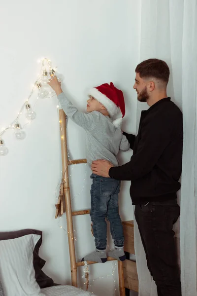 Child decorating room with his father by lights standing on stairs. Family together decorates the house for traditional celebrating winter Christmas holidays. New Year magic fairytale vibes. Vertical