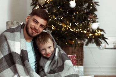 Baba oğul, Noel ağacının yanında kış tatili için süslü bir odada battaniyeye sarılmış. Aile şenlik portreleri. Yeni yıl kutlamaları. Kış büyüsü zamanı, peri masalı heyecanı.