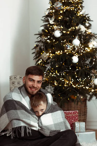 Baba oğul, Noel ağacının yanında kış tatili için süslü bir odada battaniyeye sarılmış. Aile şenlik portreleri. Yeni yıl kutlamaları. Kış büyüsü zamanı, peri masalı heyecanı. Dikey