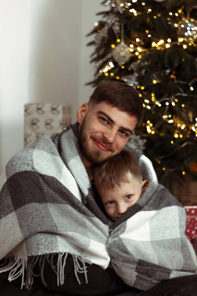 Baba oğul, Noel ağacının yanında kış tatili için süslü bir odada battaniyeye sarılmış. Aile şenlik portreleri. Yeni yıl kutlamaları. Kış büyüsü zamanı, peri masalı heyecanı. Dikey
