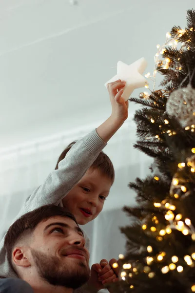 Noel ağacını süsleyen çocuk babasının omuzlarında oturuyor. Geleneksel aile, kış tatillerini kutlamak için evi dekore eder. Mutlu yüzler, sihirli peri masalı titreşimleri. Dikey