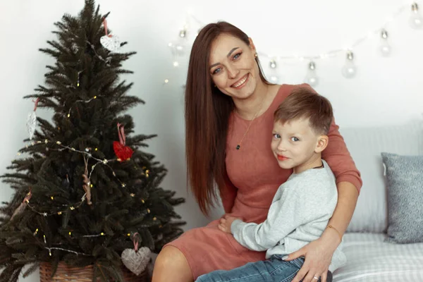 Kış tatili için süslü bir odada Noel ağacının yanında gülümseyen, sarılan ve oturan bir anne ile anne. Aile şenlik portreleri. Yeni yıl kutlamaları. Kış büyüsü zamanı, peri masalı heyecanı.