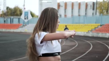 Spor giyimli genç bir bayan spor stadyumunun arka planında kollarını esnetiyor.