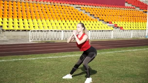 Молода струнка біла блондинка в спортивному одязі робить присідання вправи на стадіоні — стокове відео