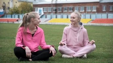 Pembe spor kıyafetleri içinde gülümseyen iki genç nilüfer çiçekli çim tarlasında oturuyorlar.