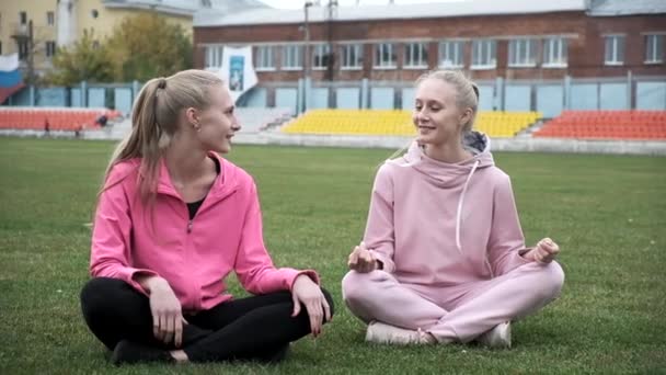 Dos jóvenes gemelos sonrientes en ropa deportiva rosa se sientan en el campo de hierba en pose de loto — Vídeo de stock