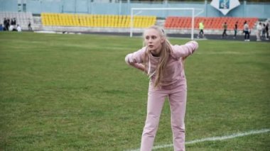 Spor kıyafetleri içinde formda bir kadın. Stadyum sahasında kollarını beline dayayıp esniyor.