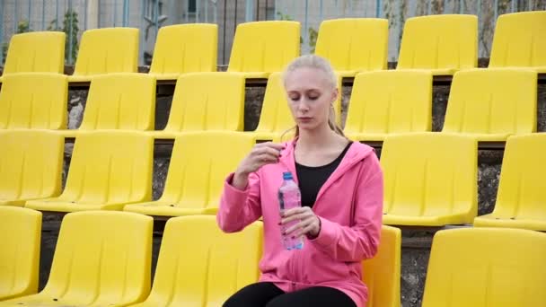 Esportivo bonita fêmea senta-se em staduim tribune, bebe água de garrafa de plástico — Vídeo de Stock
