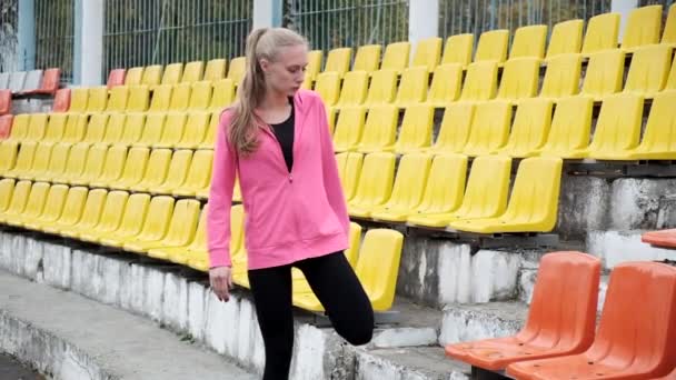 Junge, fitte Kaukasierin in Sportkleidung, die Beine auf der Tribüne des Stadions streckt — Stockvideo