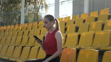 Kulaklıklı genç beyaz kadın stadyumda akıllı telefondan müzik dinliyor.