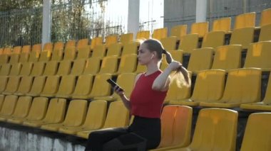 Kulaklıklı genç beyaz kadın stadyumda akıllı telefondan müzik dinliyor.