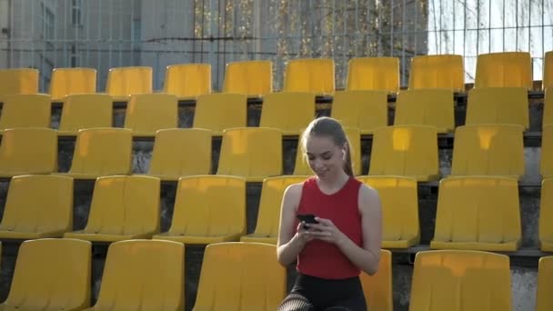 Młoda kaukaska kobieta ze słuchawkami słucha muzyki na smartfonie na stadionie — Wideo stockowe