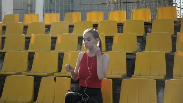 Joven mujer caucásica con auriculares escucha música en el teléfono inteligente en el estadio — Vídeo de stock