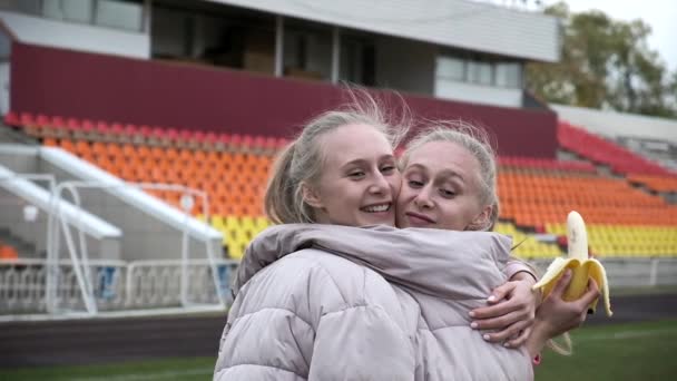 Две милые одинаковые сестры-близнецы в розовой куртке обнимаются и веселятся на стадионе — стоковое видео
